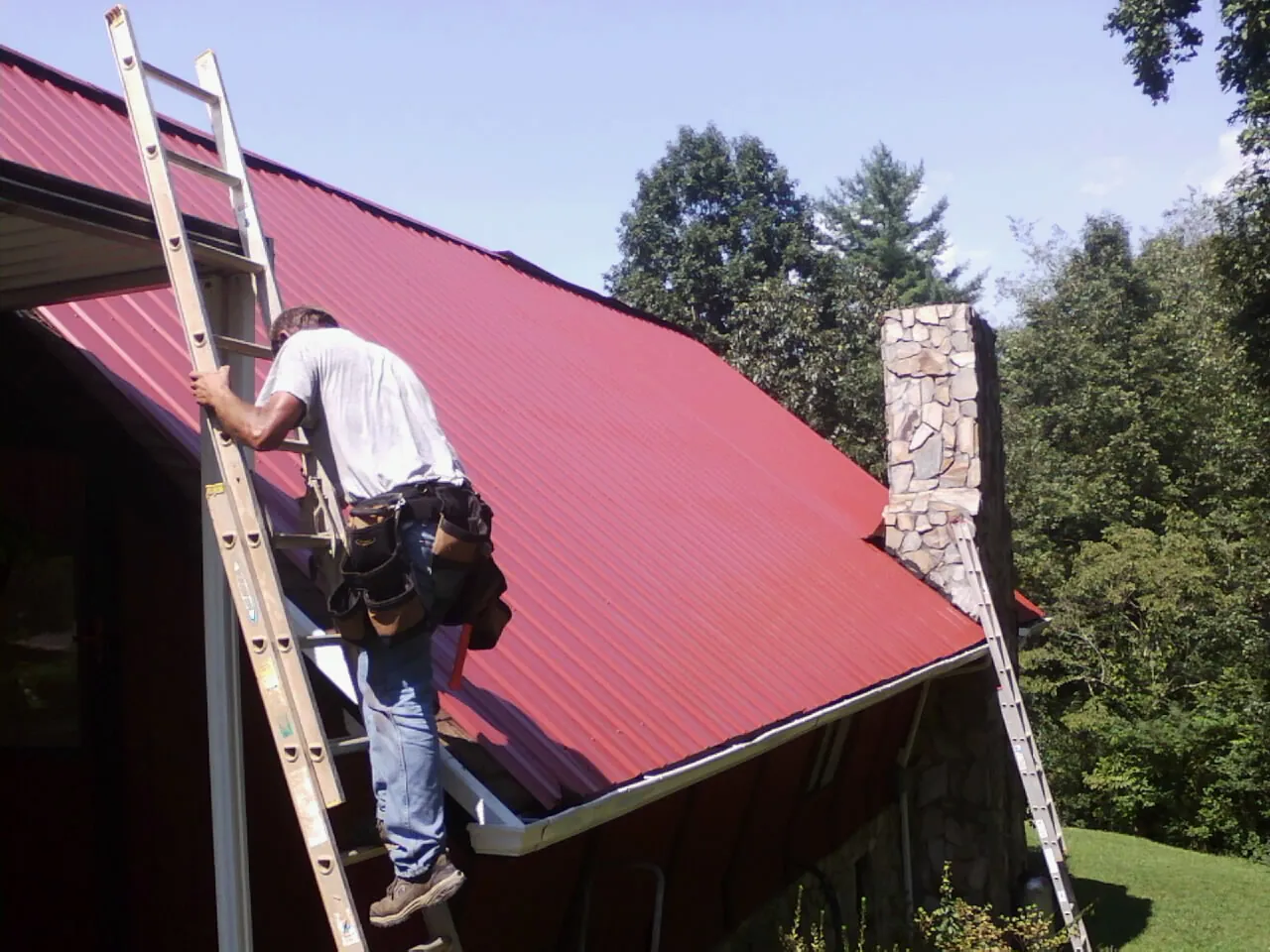 Roofer on a ladder beside a red metal roof and stone chimney — Soffit & Fascia Repair by Roofing Brothers in Andreas